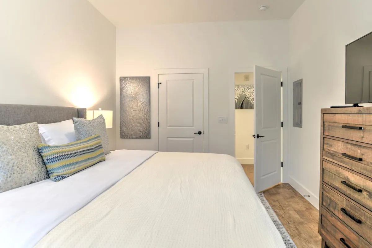 This image showcases a comfortable bed with crisp linens and decorative pillows, a modern dresser, tranquil wall art, and a television. The serene atmosphere is highlighted by soft natural light, creating an inviting space perfect for rest and relaxation.