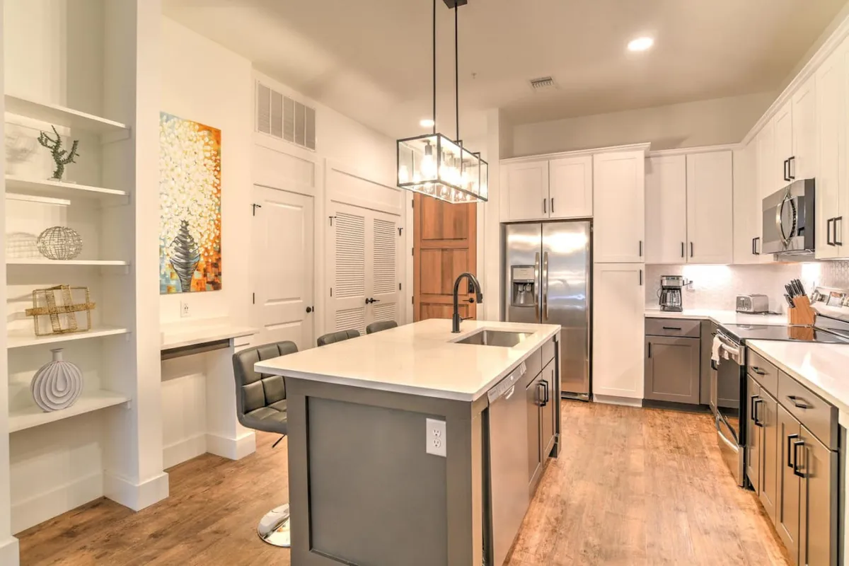 A stylish, contemporary kitchen with a spacious island at its center, illuminated by elegant pendant lighting. Stainless steel appliances line the walls, while eye-catching artwork adds a touch of personality. The open layout and bar-style seating encourage relaxed gatherings and social interaction.