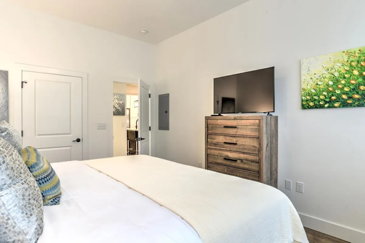 This tranquil bedroom scene features a neatly made bed with decorative pillows, a television on a modern dresser, and colorful artwork adding personality to the walls. The soft lighting and thoughtful arrangement create a peaceful, inviting space.