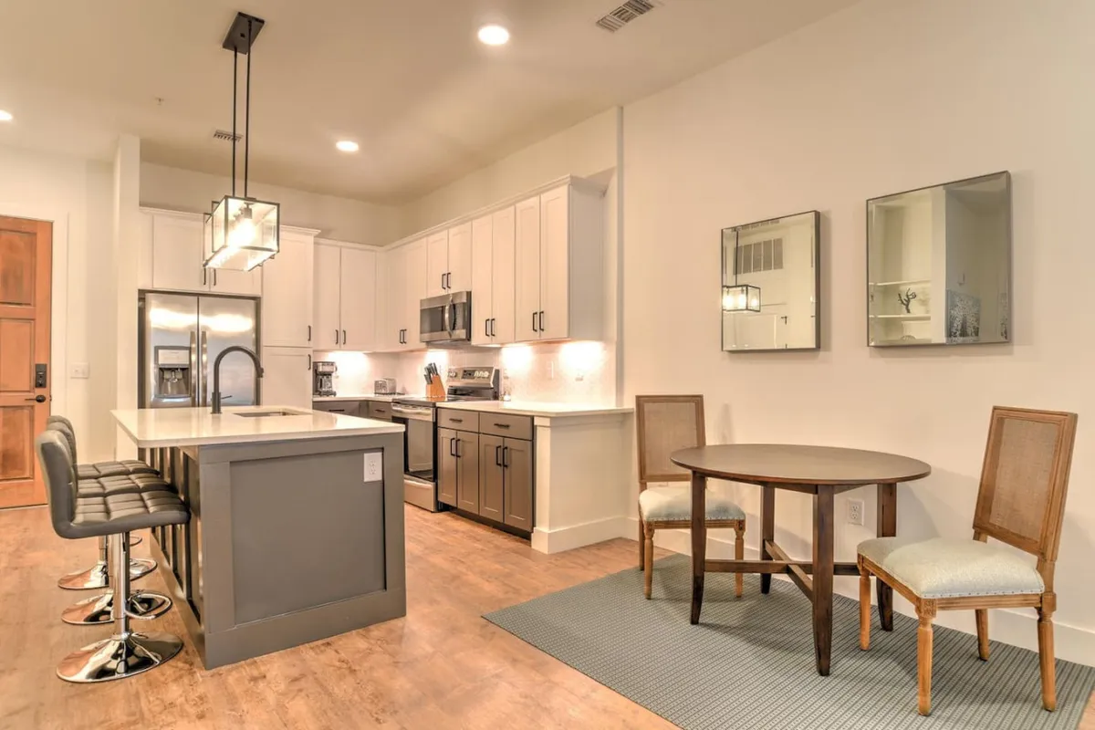 This contemporary interior highlights a kitchen island, dining table, stools, and mirrors—showcasing an open space just inside the residence. The inviting perspective hints at the transition from entry to living space, reinforcing the theme of seamless arrival.